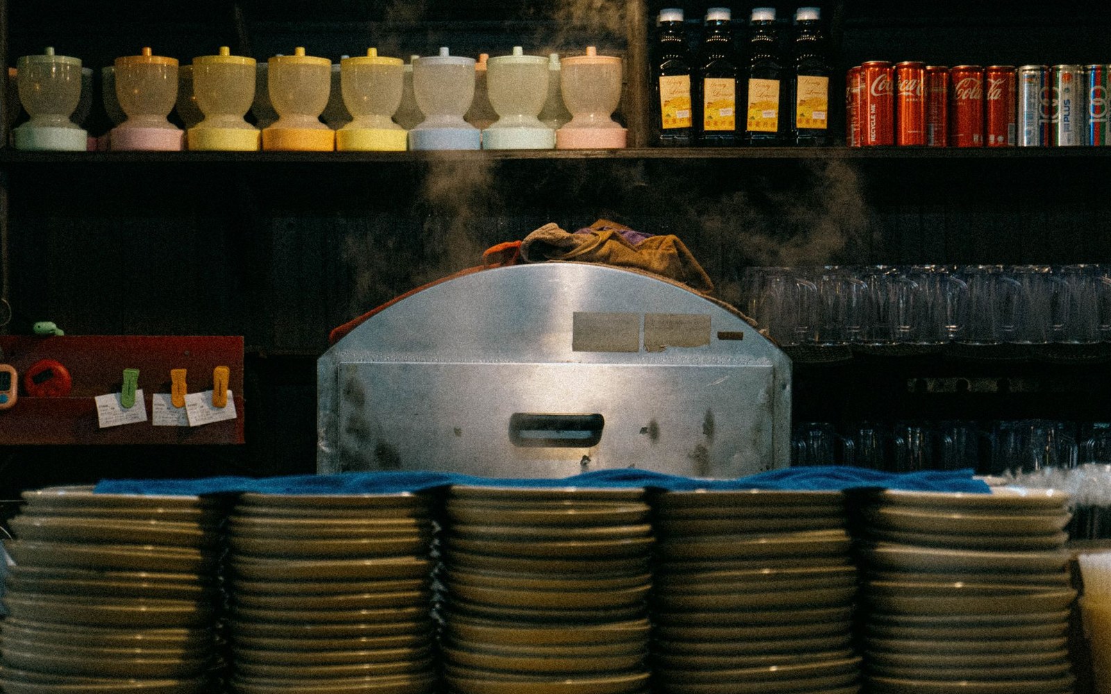 Steam rises from a metal dish warmer behind stacked plates and shelves of drinks
