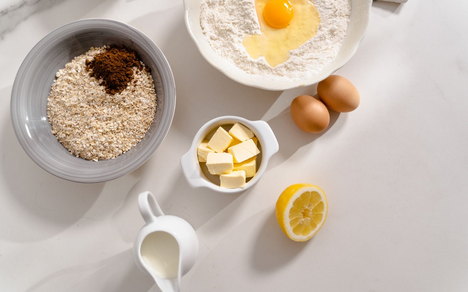 Bowls of oats, flour with a cracked egg, butter cubes, eggs, lemon and milk