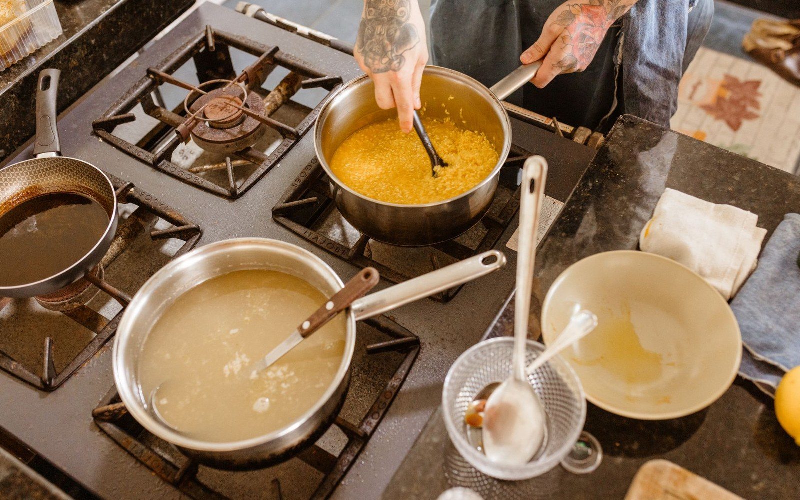 A tattooed cook stirs simmering stock in pots on a gas stove