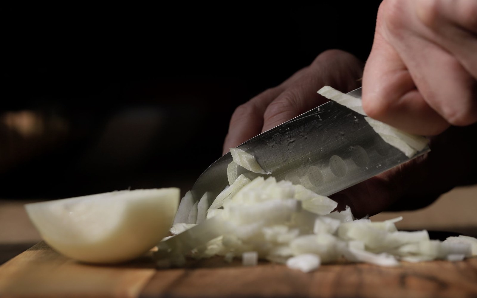 A knife blade mid-stroke through a pile of diced carrot, celery and onion on a pale wooden board